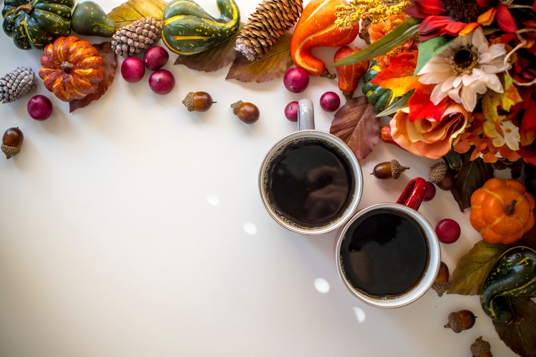 Two mugs of dark hot drinks surrounded by pumpkins, gourds, pinecones, and autumn leaves on a white surface.